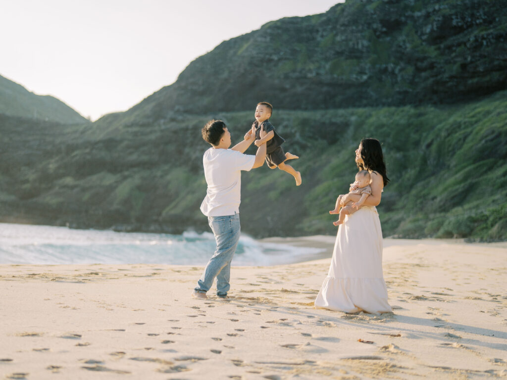 Dad lifting his laughing toddler during a family beach photo session in Hawaii while mother holds their baby.