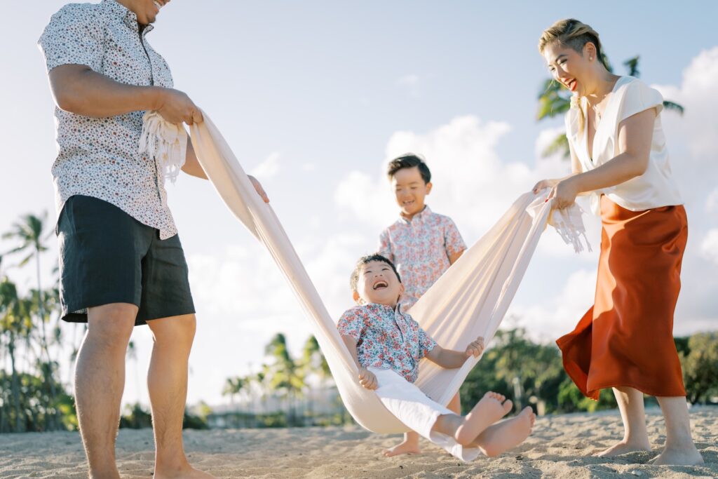 Parents laughing as they swing their young child in a blanket during a playful family beach photoshoot in Hawaii.