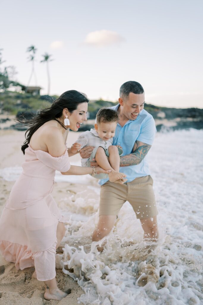 Parents laughing with their young son as waves splash around them during family beach photos in Hawaii.